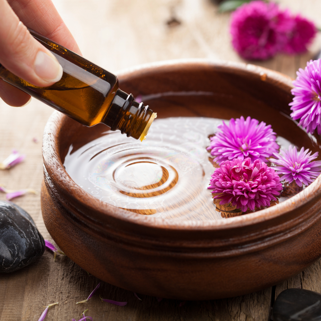 Hand pouring essential oil into a small wooden bowl, surrounded by purple flowers and spa stones, representing a calming aromatherapy ritual.
