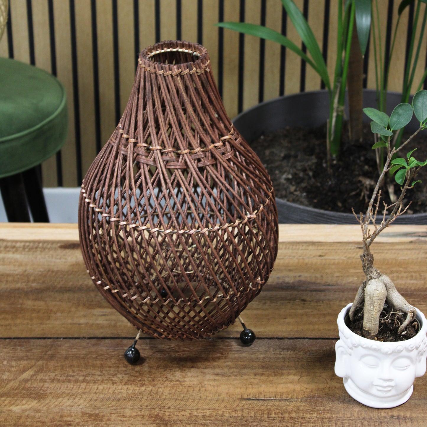Rattan lamp styled on wooden table with green plant and cup, creating a calming, earthy interior scene.