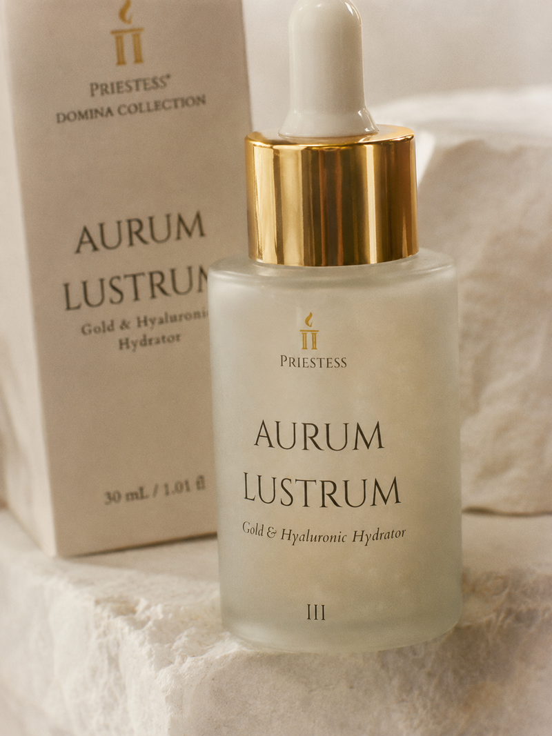 Priestess Domina Collection Aurum Lustrum Gold & Hyaluronic Hydrator serum bottle with a gold dropper cap, shown in front of its beige packaging box on a soft white textured fabric background.