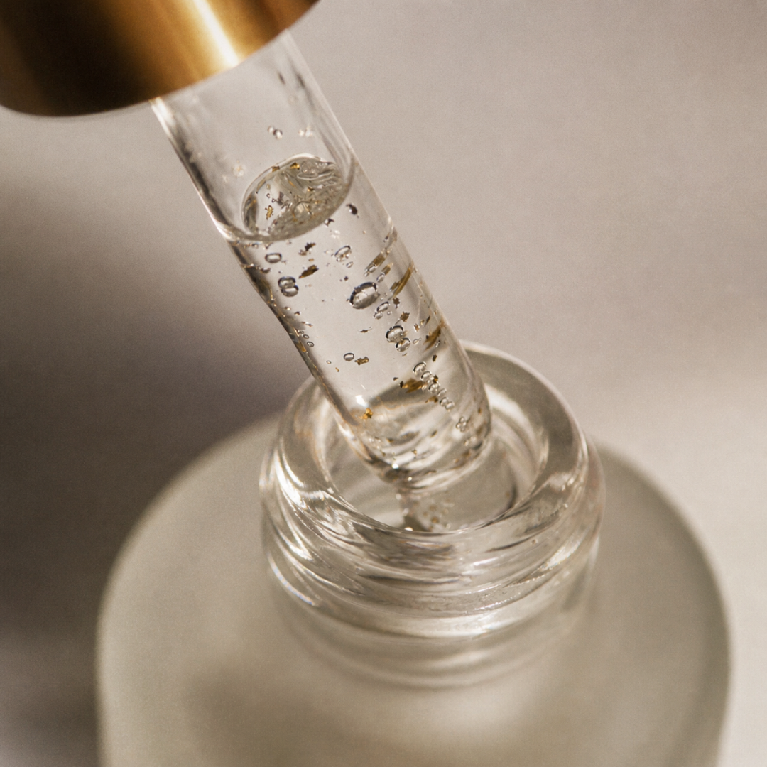 A close-up of a clear glass dropper containing a transparent gel or serum, positioned above the opening of a frosted glass bottle with a gold-toned cap, set against a soft neutral background.