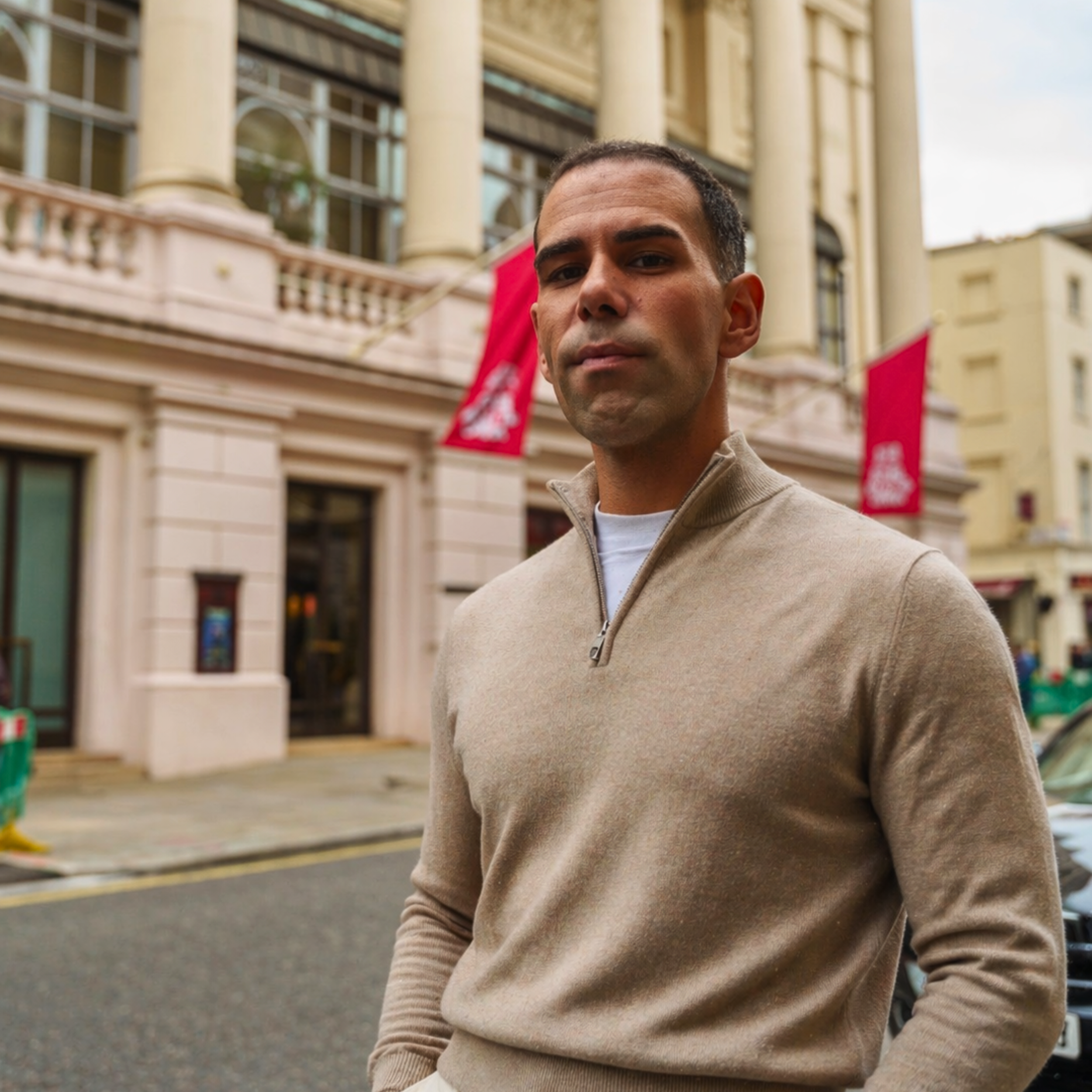 Man wearing a beige sweater standing on a city street with buildings and flags in the background