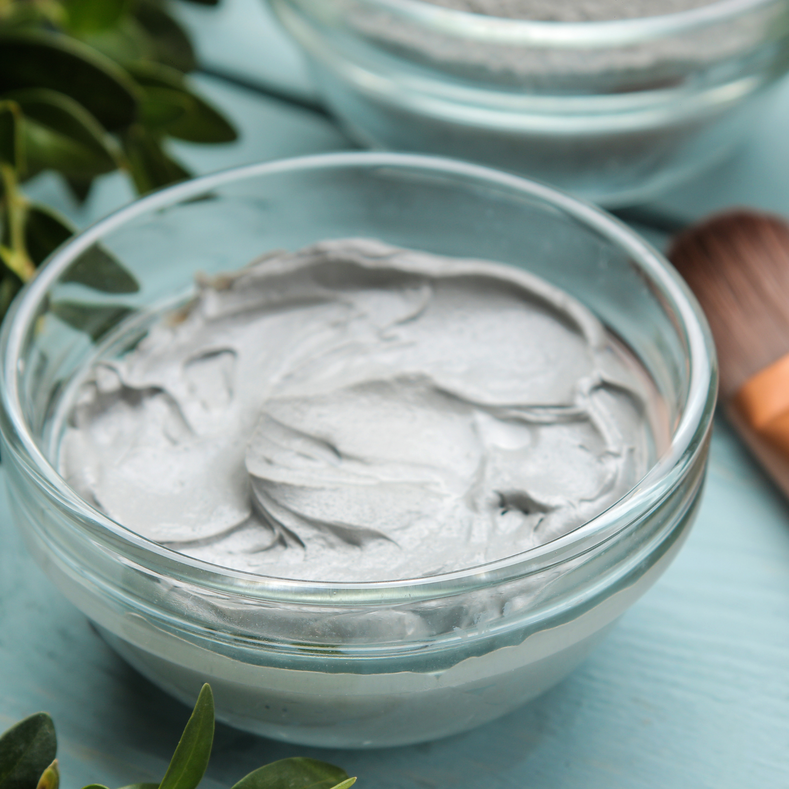 Glass bowl filled with gray cream on a light blue surface with green leaves.