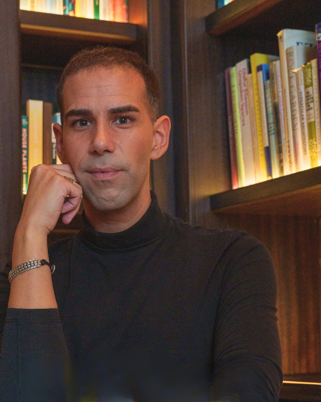 Man in a black turtleneck sitting in front of a bookshelf with books.