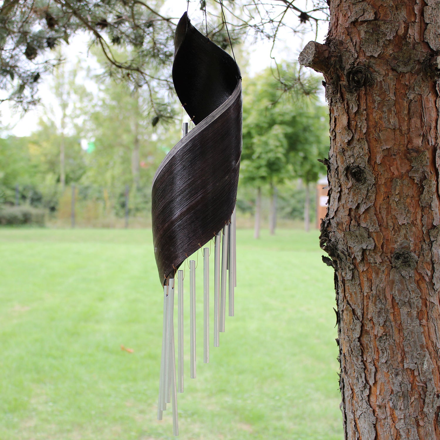 Side profile of brown coconut leaf chime with silver sound tubes hanging outdoors for tranquil ambiance.